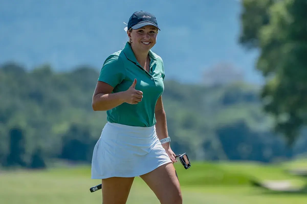 young female golfer walks with club in hand giving thumbs up