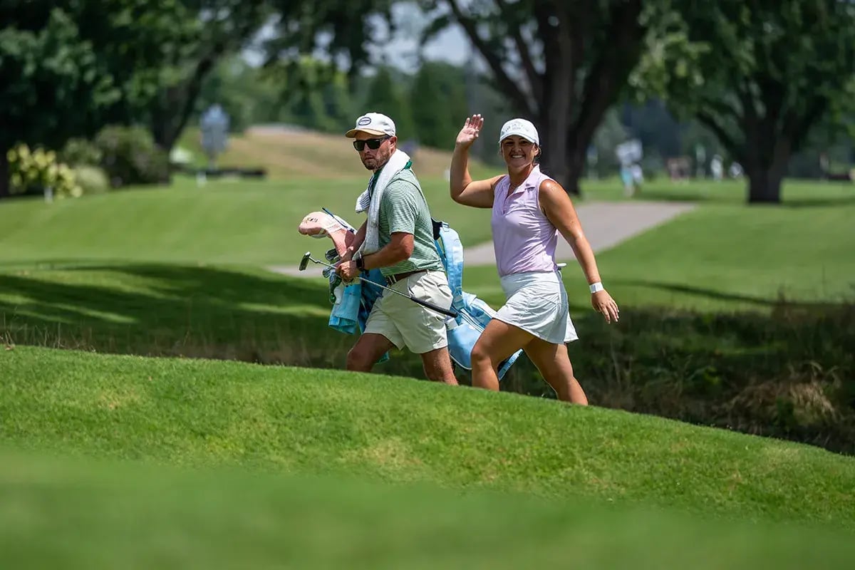 young female golfer waves smiles walking with caddie